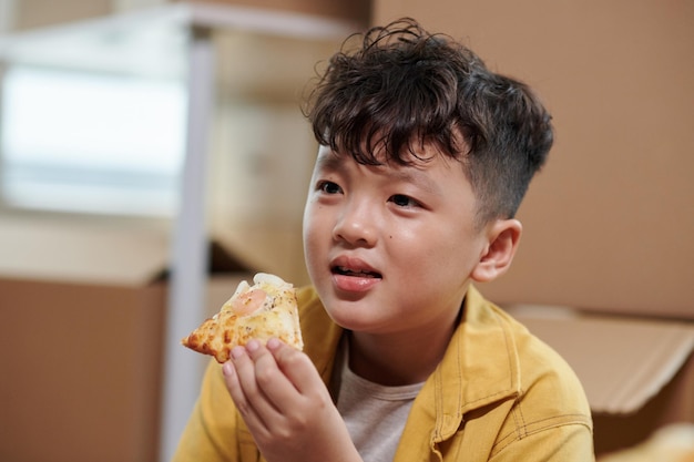 Preteen Vietnamese boy eating slice of pizza in new house