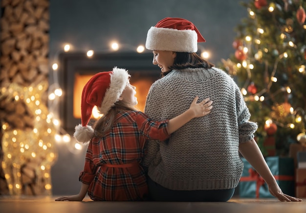 Prettige kerstdagen en fijne feestdagen. moeder en haar schattige dochtermeisje kijken naar open haard. ouder en klein kind hebben plezier in de buurt van boom binnenshuis.