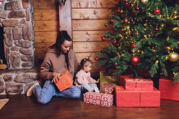 Prettige kerstdagen en fijne feestdagen Vrolijke moeder en haar schattige dochtermeisje wisselen cadeaus. Ouder en klein kind plezier in de buurt van de kerstboom binnenshuis. Liefdevolle familie met cadeautjes in de kamer.