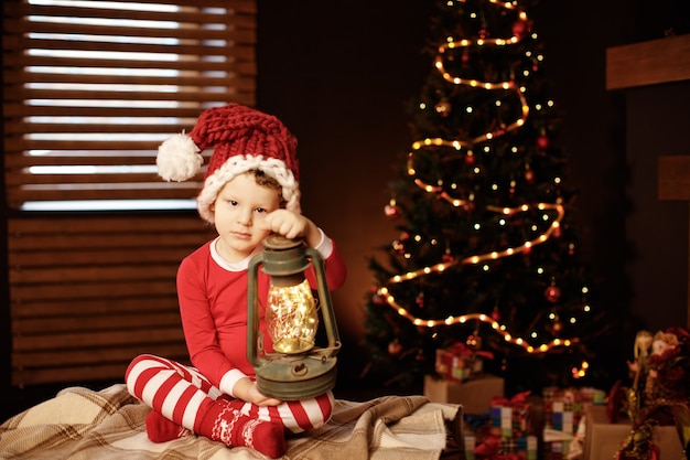 Prettige kerstdagen en prettige feestdagen Een kleine jongen zit met een lantaarn bij de kerstboom Nieuwjaar, elf.