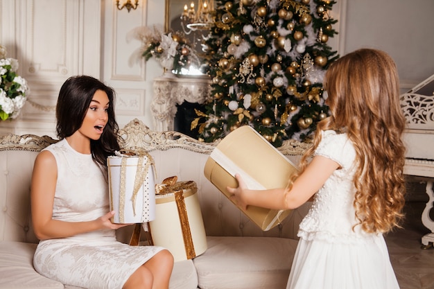 Prettige kerstdagen en prettige feestdagen. Vrolijke moeder en haar schattige dochter meisje geschenken uitwisselen in witte klassieke interieur piano en een versierde kerstboom. Nieuwjaar