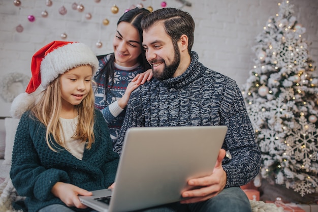 Prettige kerstdagen en prettige feestdagen Vrolijke moeder, vader en haar schattige dochter meisje om een laptop te gebruiken. Ouder en klein kind plezier binnenshuis in de buurt van de kerstboom. Morning Xmas.