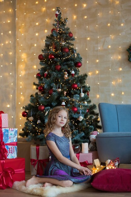예쁜 10 살짜리 아이가 화환과 선물 사이에서 크리스마스 트리 앞에 앉아 있습니다.