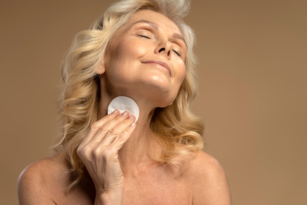 Pretty 5055 years old blonde Caucasian woman using cotton pad removing makeup on beige background