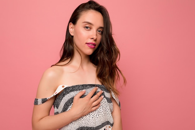 Photo pretty adorable girl with wavy hair with pink lips is posing at camera over pink background she is dressed light summer dress with bare shoulders