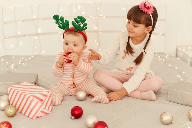 Fratelli adorabili che indossano abiti casual e cerchi da festa seduti sul letto e giocano con le palline, ragazza più anziana che guarda sua sorella con amore, felice di festeggiare il natale.