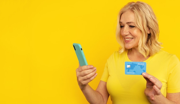 Pretty adult lady in bright clothes watching camera and posing with blue phone and credit card.