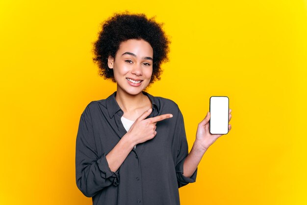 Pretty african american or brazilian woman looks at camera hold in hand smartphone with white empty mockup screen for presentation and points finger at it stand on isolated yellow backgroundsmile