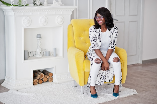 Pretty african american woman in eyeglasses posed in room, sitting on yellow chair.