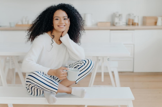 Pretty African American woman white jumper striped pants socks leisurely poses on kitchen bench with tea cup Relaxed morning coffee at cozy home