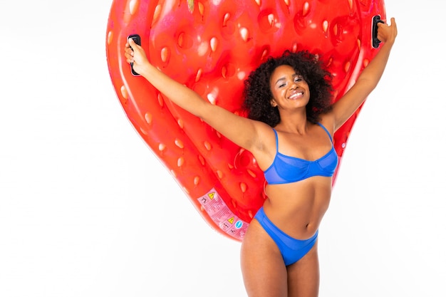 Pretty african female stands in swimsuit with rubber beach strawberry mattress and smiles isolated