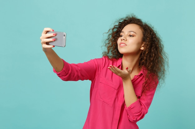 Pretty african girl in casual clothes blowing sending air kiss, doing selfie shot on mobile phone isolated on blue turquoise background. People sincere emotions, lifestyle concept. Mock up copy space.