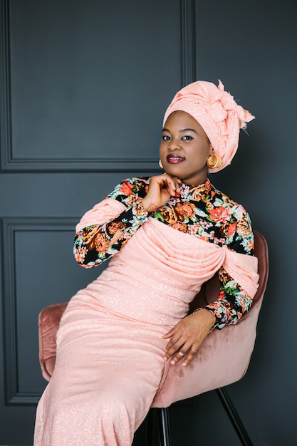 Pretty African woman wearing an pink colorful fashionable dress and headscarf, sitting on the chair on dark blue studio wall background and looking at camera with smile