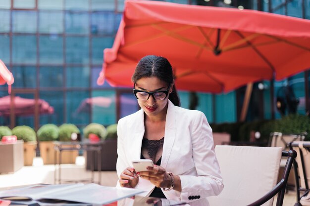 Довольно афро деловая женщина в белой куртке сидит в летнем кафе и использует смартфон