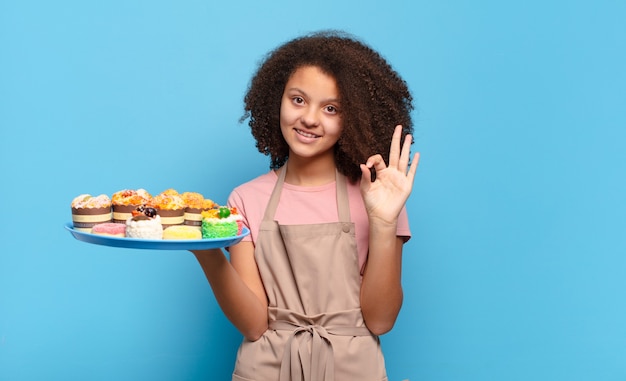 Pretty afro teenager feeling happy, relaxed and satisfied, showing approval with okay gesture, smiling. humorous baker concept