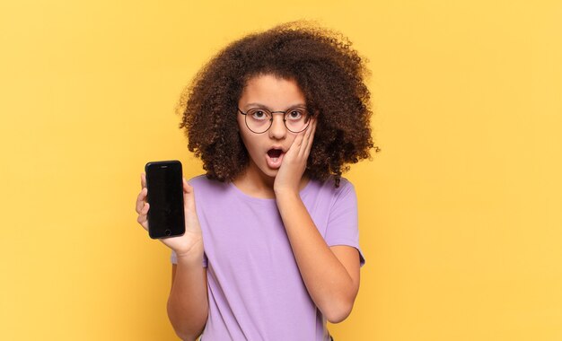 Pretty afro teenager feeling shocked and scared, looking terrified with open mouth and hands on cheeks and holding a cell