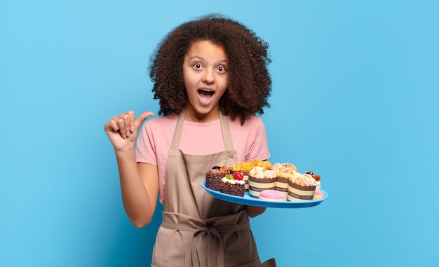 Pretty afro teenager looking astonished in disbelief, pointing at object on the side and saying wow, unbelievable. humorous baker concept