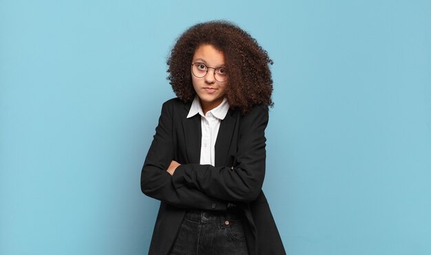 Pretty afro teenager shrugging, feeling confused and uncertain, doubting with arms crossed and puzzled look. humorous business concept