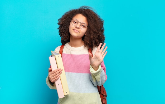 Pretty afro teenager smiling happily and cheerfully, waving hand, welcoming and greeting you, or saying goodbye. student concept