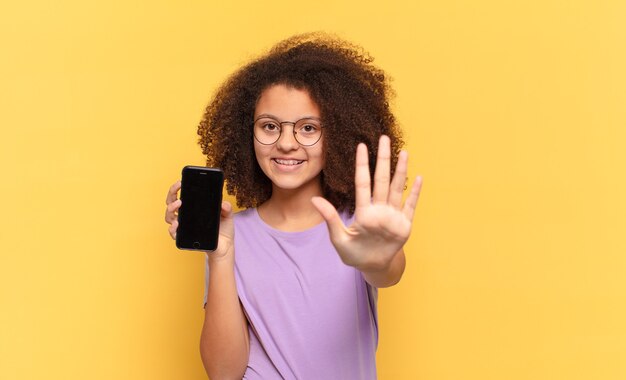 Pretty afro teenager smiling and looking friendly, showing number five or fifth with hand forward, counting down and holding a cell