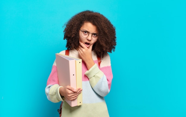 Pretty afro teenager with mouth and eyes wide open and hand on chin, feeling unpleasantly shocked, saying what or wow. student concept