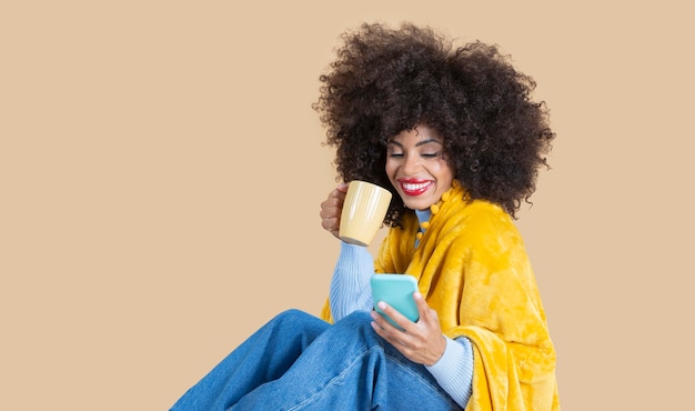 Pretty afro woman covered with a blanket while having a coffee\
and looking at her smarpthone