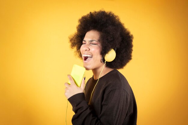 ヘッドフォンとスマートフォン、黄色の背景で音楽を聴いているかなりアフロの女性