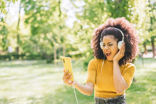 Pretty afro woman listens to music with headphones dancing in a garden. Selective focus.