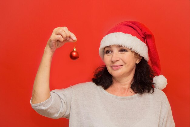 Donna abbastanza invecchiata in cappello da babbo natale che tiene la palla di natale nelle sue mani su sfondo rosso