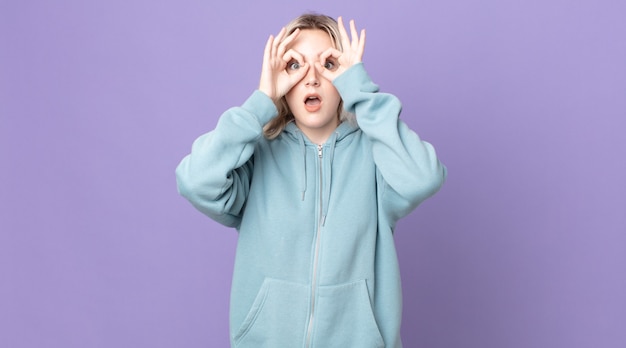 Pretty albino woman feeling shocked, amazed and surprised, holding glasses with astonished, disbelieving look