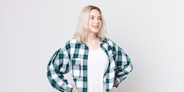 Pretty albino woman looking happy, cheerful and confident, smiling proudly and looking to side with both hands on hips