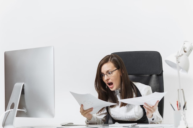 Pretty angry business woman in suit sitting at the desk with documents, working at computer with modern monitor in light office, swearing and screaming, resolving issues,