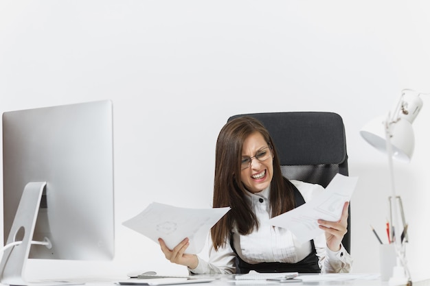 Pretty angry business woman in suit sitting at the desk with documents, working at computer with modern monitor in light office, swearing and screaming, resolving issues,