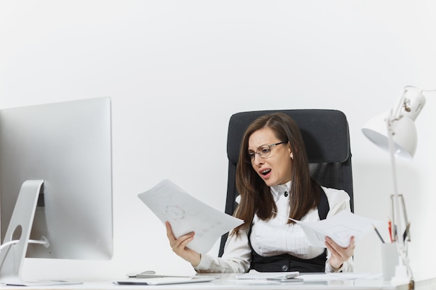 Pretty angry business woman in suit sitting at the desk with documents, working at computer with modern monitor in light office, swearing and screaming, resolving issues,