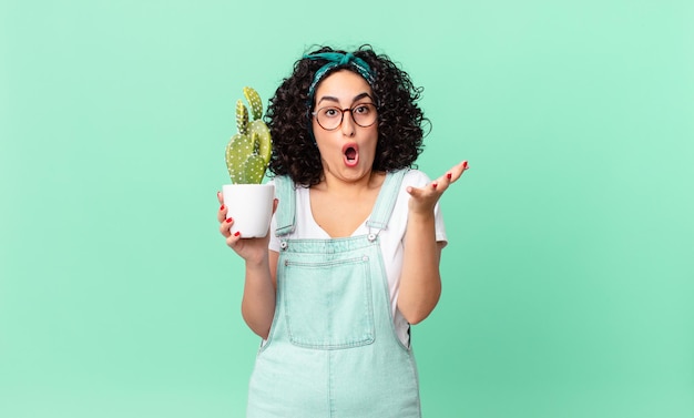 Pretty arab woman amazed, shocked and astonished with an unbelievable surprise and holding a potted cactus