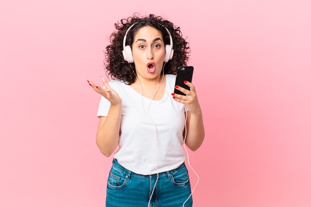 Pretty arab woman amazed, shocked and astonished with an unbelievable surprise with headphones and a smartphone