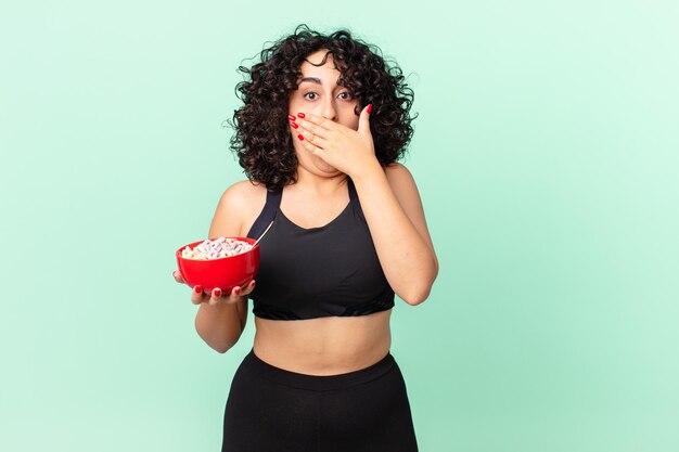 Pretty arab woman covering mouth with hands with a shocked wearing fitness clothes and holding a corn flakes bowl