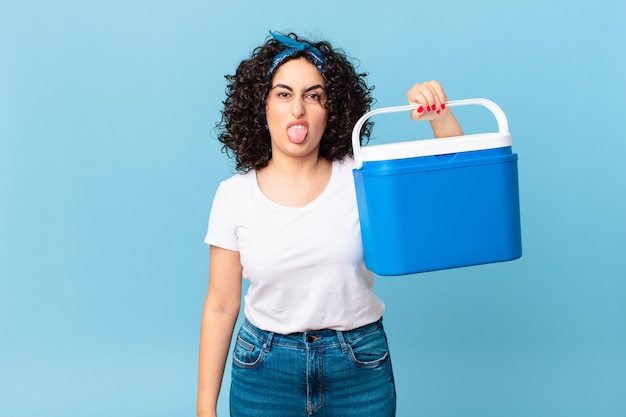 Pretty arab woman feeling disgusted and irritated and tongue out and holding a portable refrigerator