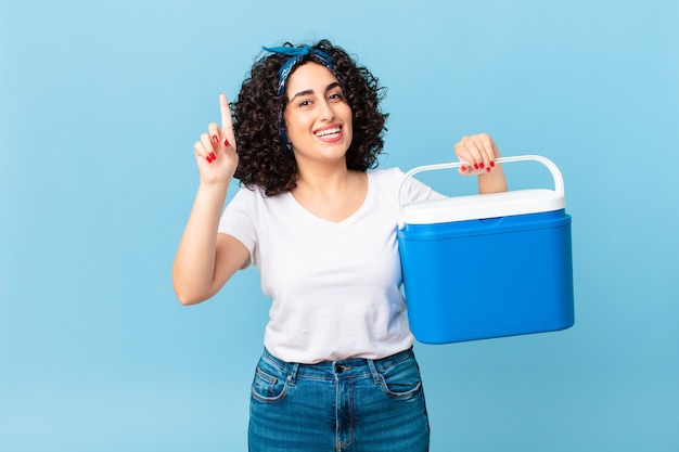 Pretty arab woman feeling like a happy and excited genius after realizing an idea and holding a portable refrigerator