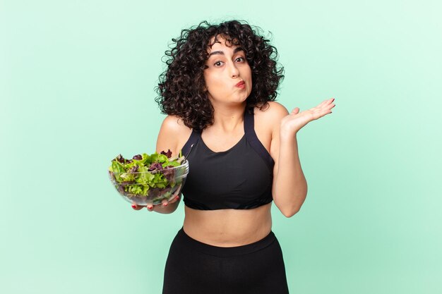 Foto donna abbastanza araba che si sente perplessa e confusa e dubita e tiene in mano un concetto di dieta a base di insalata