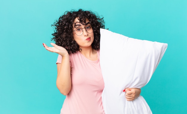 Foto bella donna araba che si sente perplessa, confusa e dubbiosa. in pigiama e con in mano un cuscino