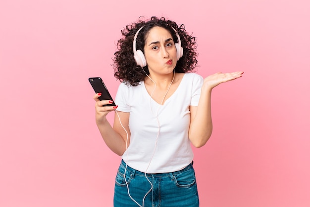 Pretty arab woman feeling puzzled and confused and doubting with headphones and a smartphone