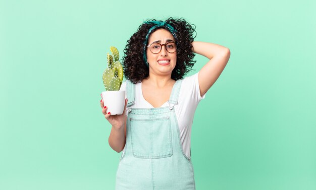 Pretty arab woman feeling stressed, anxious or scared, with hands on head and holding a potted cactus