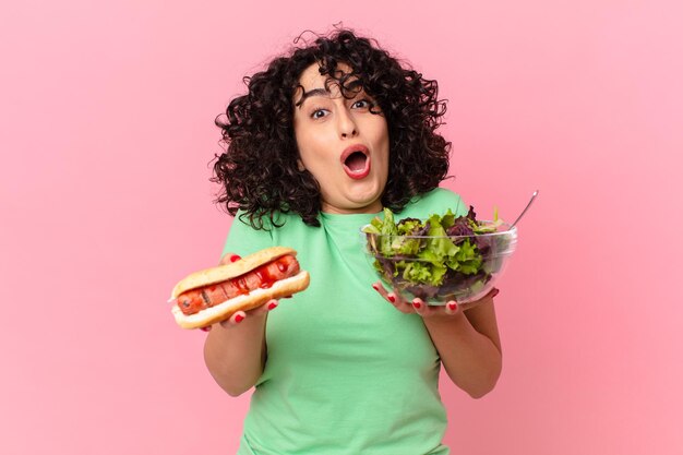 Pretty arab woman holding a salad and a hot dog. diet concept