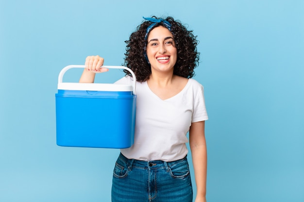 Pretty arab woman looking happy and pleasantly surprised and holding a portable refrigerator