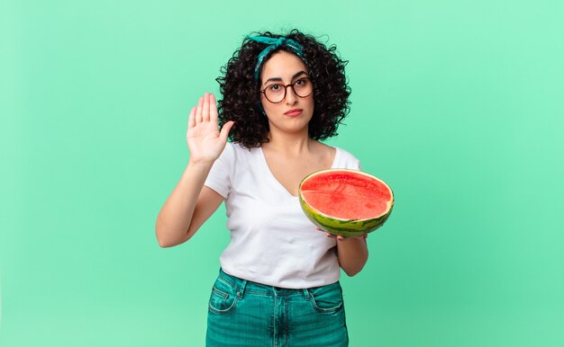 Pretty arab woman looking serious showing open palm making stop gesture and holding a watermelon. summer concept