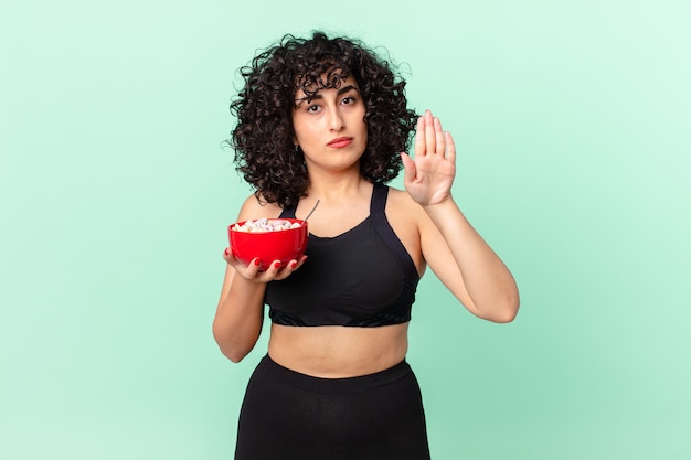 Pretty arab woman looking serious showing open palm making stop gesture wearing fitness clothes and holding a corn flakes bowl