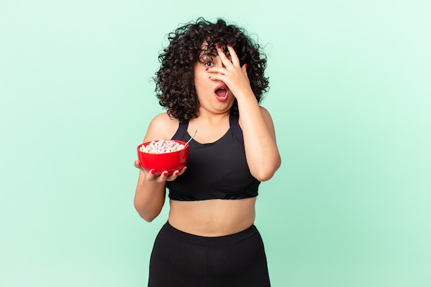 Photo pretty arab woman looking shocked, scared or terrified, covering face with hand wearing fitness clothes and holding a corn flakes bowl