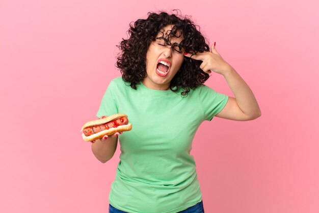 Pretty arab woman looking unhappy and stressed suicide gesture making gun sign and holding a hot dog