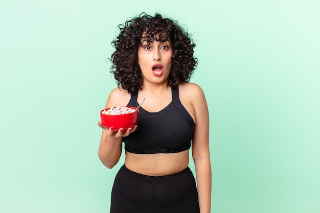 pretty arab woman looking very shocked or surprised wearing fitness clothes and holding a corn flakes bowl